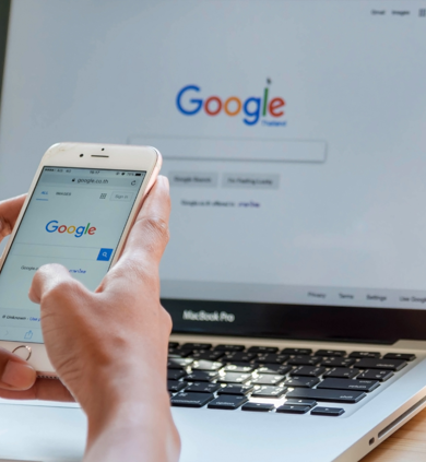 Person using Google Search on mobile phone with Google on desktop in background