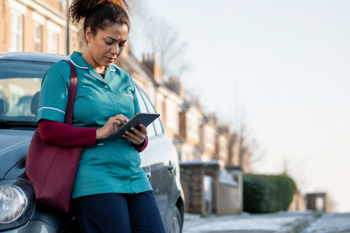 A home care worker holding a mobile device. She is standing outside after her visit, leaning against her car, taking visit notes on her tablet.