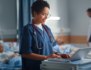 A nurse on a laptop whilst an old lady is lying down on a hospital bed