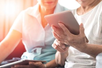 an elderly person and a younger lady sitting on a couch while looking at the screen of a tablet