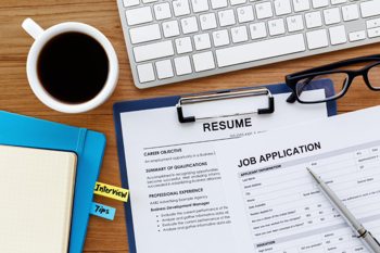Image depicts a keyboard, a coffee cup, a pair of glasses, notebooks, and job application files on a table