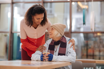 Young woman standing beside an older lady seated at a table, placing a hand on the older adult’s shoulder while the older lady holds a glass. They are indoors, with blurred windows and décor in the background.