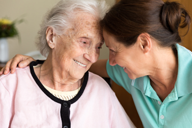 A nurse who has her arm around an elderly woman, both smiling 