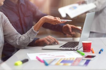 Close-up of two people sitting at a desk in front of a laptop; on the desk there is a cup of coffee, pens and papers.