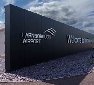 Entrance sign at Farnborough Airport with the words 'Farnborough Airport' and 'Welcome to Farnborough' on a black wall under a cloudy sky