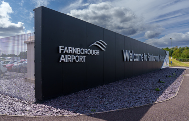 Entrance sign at Farnborough Airport with the words 'Farnborough Airport' and 'Welcome to Farnborough' on a black wall under a cloudy sky