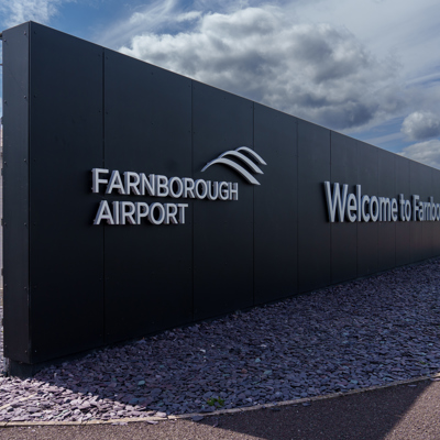 Entrance sign at Farnborough Airport with the words 'Farnborough Airport' and 'Welcome to Farnborough' on a black wall under a cloudy sky
