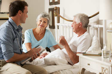 eldermy man lying down in a bed, with a woman and a male care worker beside him