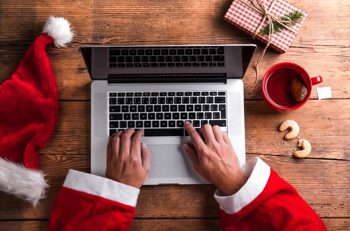 Person wearing a Santa Claus outfit typing on a laptop, with a Santa hat, wrapped gift, cup of tea, and Christmas cookies nearby.