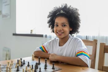 A young girl playing chess at home pauses to have her photograph taken by a relative.