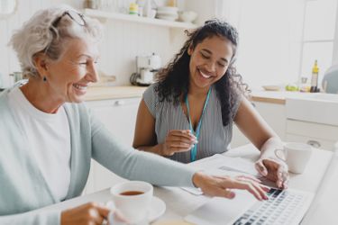 A female care worker sitting together with an elderly lady at a kitchen table, looking at the screen of a laptop.