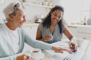 A female care worker sitting together with an elderly lady at a kitchen table, looking at the screen of a laptop.