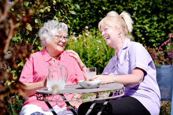 female care worker sitting together with an elderly lady in the garden of a care home; on the table in front of them there is a jug of water and a bowl of strawberries