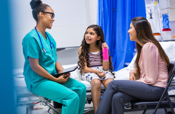 Nurse sitting on the bed with a young child who's arm is broken and her mother sitting down on a chair besides her smiling