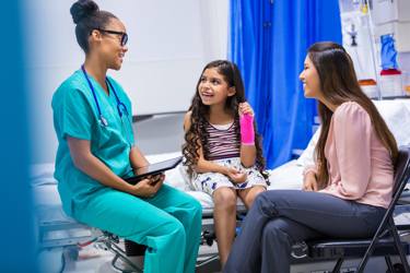 Nurse sitting on the bed with a young child who's arm is broken and her mother sitting down on a chair besides her smiling