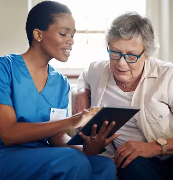 A black nurse showing something to an elderly lady on the tablet 