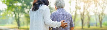 care worker holding their hand against an elderly lady's back while they walk together in a park