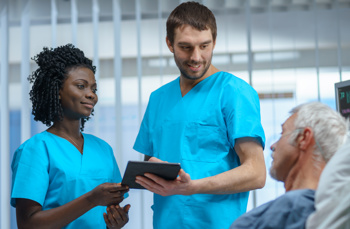 A black woman and a white male consulting a unwell elderly man who is laying in a hospital bed