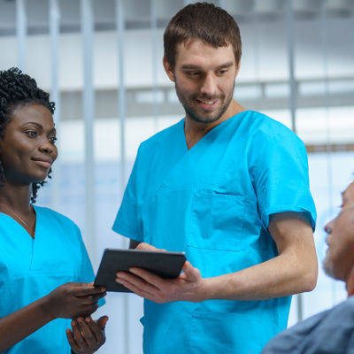 A black woman and a white male consulting a unwell elderly man who is laying in a hospital bed
