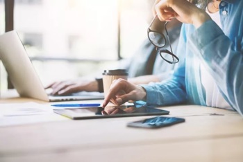 a young person sitting at a desk holding a pair of glasses and scrolling on a tablet