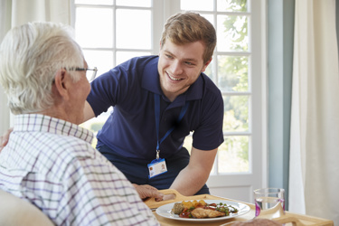 male care worker checking on an elderly resident while he serves his meal