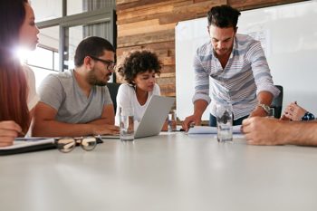 four young people sitting around a desk, with laptops and notepads, having a discussion