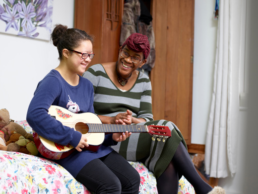 a little girl with special needs playing the guitar, whilst a black woman is looking at her smiling 