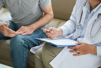 Close-up of a healthcare worker and a man sitting on a couch. The worker has a file and a pen in their hand.