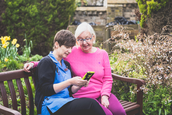 care worker and elderly resident sitting together on a bench in a garden while looking at the screen of a phone