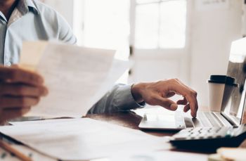 Person typing on a laptop while holding papers at a desk with office supplies