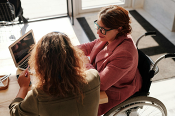 Two young women, one of them in a wheelchair, sitting at a desk in a brightly illuminated room, looking at the screen of a laptop.