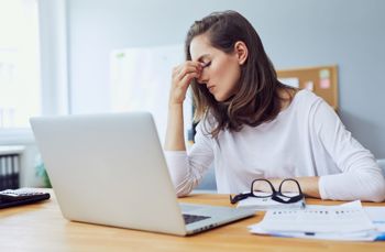 Person holding their forehead at a desk with charts and glasses, suggesting stress or fatigue