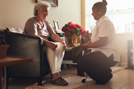A nurse kneeling down talking to an elderly woman sitting on a chair smiling 