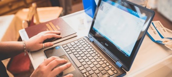 image depicts a laptop on a table and a pair of hands typing on the keyboard