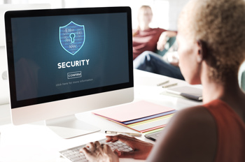 A young person sitting in front of a computer with their hands on the keyboard. The computer's screen depicts a blue shield with a keyhole in it, below it reads'security'.