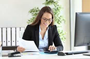 Person in a blazer using a calculator at a desk with documents and a computer monitor