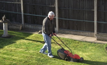 An elderly man, Neville, mowing his back garden lawn.