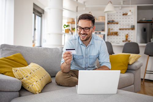 Man sat on sofa smiling at payment card with laptop in front of him