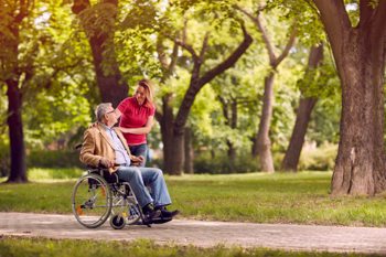 care worker and an elderly man in a wheelchair taking a stroll through a park