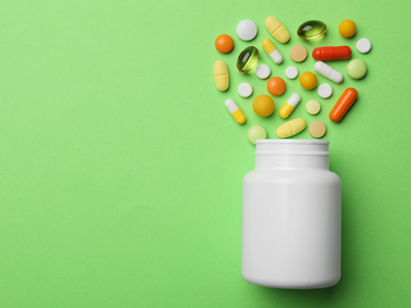 A pill bottle with several white, yellow and orange pills and capsules coming out of it, against a green background