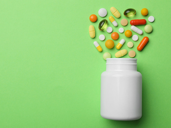 A pill bottle with several white, yellow and orange pills and capsules coming out of it, against a green background