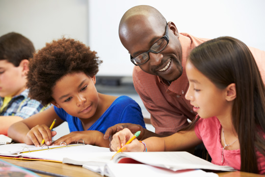 A black male teacher helping two young girls with their work whilst smiling 