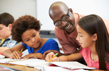A black male teacher helping two young girls with their work whilst smiling