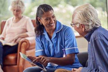 Care worker sitting together with an elderly man on a couch in a care home. The Care worker is showing the elderly man something on the screen of her tablet.