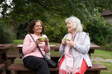 two elderly ladies sitting outside on a bench, having a cup of tea