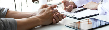 close-up of two people sitting at a desk during a conversation, with one person’s hands clasped and the other holding a pen near a clipboard and documents