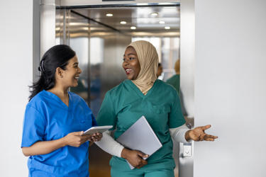A black female nurse with a hijab and an Asian female nurse smiling at each other