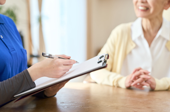 Care worker sitting at a table with an elderly lady. The care worker has a file and a pen in her hand.
