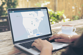 Close-up of a person typing on a laptop. The screen of the laptop is displaying a map with location pins. Beside the laptop there is a coffee cup.